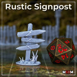 Photograph of a printed Rustic Signpost terrain piece by Mia Kay M3DM, unpainted in grey resin, shown on a gridded gaming mat beside a large bronze d20 for scale. The piece shows multiple rope-lashed wooden boards with carved markings on a textured base with grass tufts.