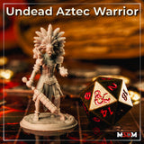 Photograph of a printed and unpainted Undead Aztec Warrior miniature in grey resin, showing the full sculpt with feathered headdress, ceremonial armour, macuahuitl, and detailed base, photographed beside a large D&D d20 dice on a tabletop gaming mat