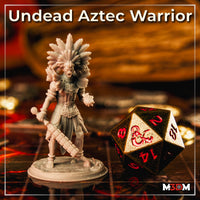 Photograph of a printed and unpainted Undead Aztec Warrior miniature in grey resin, showing the full sculpt with feathered headdress, ceremonial armour, macuahuitl, and detailed base, photographed beside a large D&D d20 dice on a tabletop gaming mat