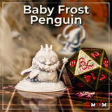Photograph of the printed Baby Frost Penguin miniature by Mia Kay M3DM — an unpainted white resin mini shown next to a large gold and red metal d20 on a tabletop gaming mat, showing the full sculpt with its ice-spike crown, expressive face, textured feathers, and star-headed staff