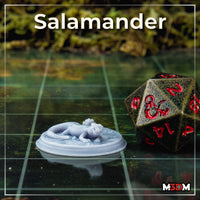 Printed and unpainted resin Salamander miniature on a gaming mat next to a large D20 die, showing the actual print scale — the small chibi salamander rests on its detailed lily pad base with lily buds sculpted around it