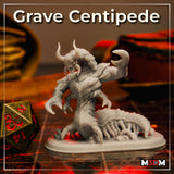Unpainted grey resin print of the Grave Centipede miniature on a tabletop gaming mat beside a large gold D20 dice — showing the full sculpt with horned skull head, open jaw, raised claws, and detailed segmented centipede body