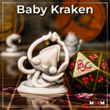 Photograph of an unpainted grey resin baby kraken miniature on a tabletop gaming mat, shown next to a large gold and red d20 for scale reference, with other unpainted miniatures visible in the background — chibi kraken sculpt by Mia Kay M3DM