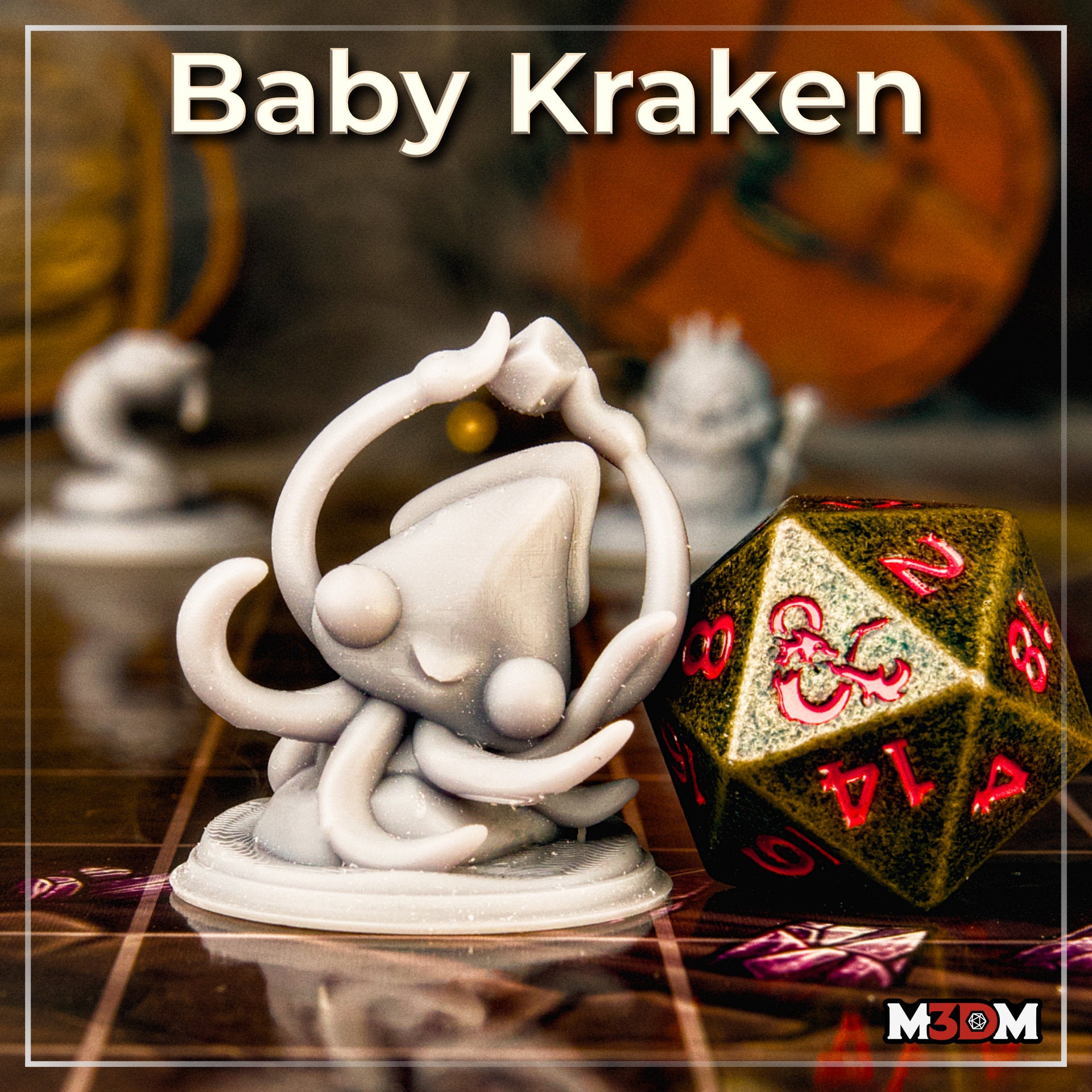 Photograph of an unpainted grey resin baby kraken miniature on a tabletop gaming mat, shown next to a large gold and red d20 for scale reference, with other unpainted miniatures visible in the background — chibi kraken sculpt by Mia Kay M3DM