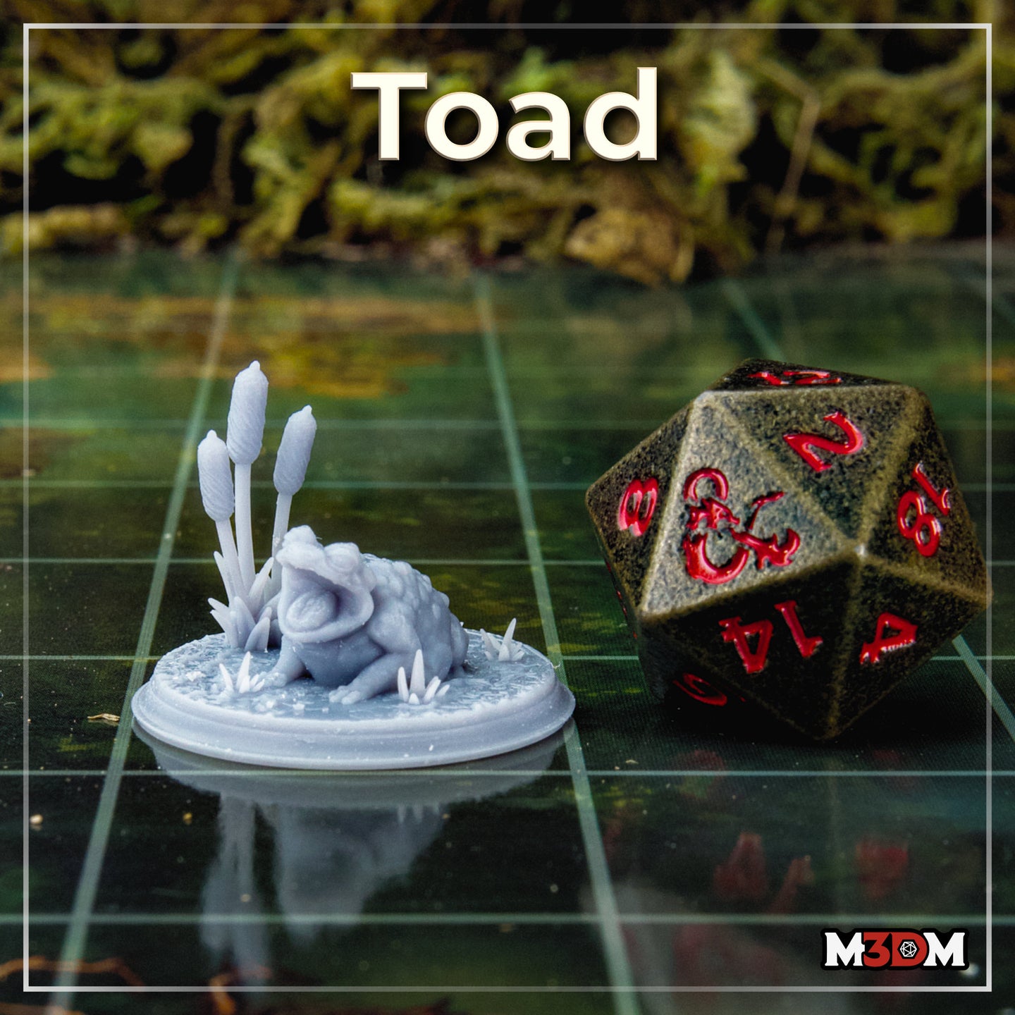 Unpainted grey resin toad miniature by Mia Kay M3DM on a gaming mat beside a D20 die, showing the open-mouthed pose on a scenic base with bulrush stalks, giving a sense of the 32mm scale