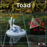 Unpainted grey resin toad miniature by Mia Kay M3DM on a gaming mat beside a D20 die, showing the open-mouthed pose on a scenic base with bulrush stalks, giving a sense of the 32mm scale