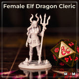 Female elf dragon cleric miniature photographed on a gaming table beside a large d20. Tall horned trident staff, long hair, wrapped bracers, layered tunic with arcane swirl detailing. Unpainted grey resin. Sculpt by Mia Kay M3DM.