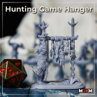 Grey resin print of the Hunting Game Hanger by Mia Kay M3DM on a gaming table — two rope-bound poles with a crossbar hung with pelts and feathered bundles, beside a d20 die for scale.