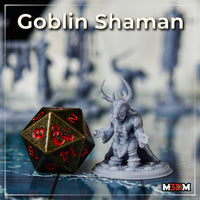 Goblin Shaman miniature by Mia Kay M3DM shown at gaming table scale beside a large D20 die, grey resin unpainted, antlered headdress and raised clawed hands visible, textured fur cloak and rope-wrapped robes clearly detailed.