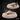 Two digital render views of an unpainted resin platypus miniature on scenic oval bases with rippled water and marsh reeds — front-facing variant in foreground, rear-angled variant behind, shown against a dark textured background