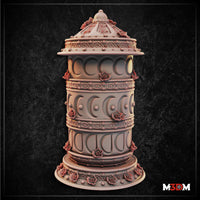 Unpainted white resin prayer wheel terrain piece shown beside a large d20 die for scale, displaying the full tiered column, domed top, and cherry blossom detailing across the ornate carved body. Sculpt by Mia Kay M3DM.