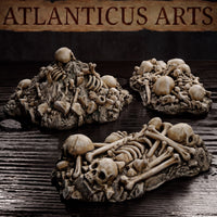 Close view of three Atlanticus Arts bone pile terrain pieces on a dark surface — mounded skull heaps with layered bones and a flat elongated piece showing multiple skeletal forms with visible ribcages and cranial detail.