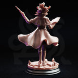 Rear view of Professor Margaux de la Croix miniature — showing the dramatic sweep of her quilted cloak, belted waist, and the open tome clutched in her left hand, with writhing hair detail visible from behind