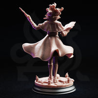 Rear view of Professor Margaux de la Croix miniature — showing the dramatic sweep of her quilted cloak, belted waist, and the open tome clutched in her left hand, with writhing hair detail visible from behind