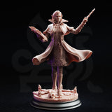 Front view of unpainted grey resin miniature of Professor Margaux de la Croix — elven wizard in ornate long coat with scrollwork trim, cloak swept to one side, wand raised, spellbook open, standing on a scenic base with melted candles and a skull