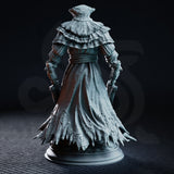Imposing robed figure in a tightly wound hood carrying twin asymmetric blades — one cleaver-like, one curved — with segmented pauldrons and knotted rope sash, standing on a rubble and severed head base. Sculpt by DM Stash.