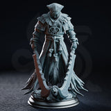 Hooded executioner in layered frayed robes holding two large curved blades, standing over a screaming severed head base, with a ceremonial bell hanging from rope-bound waist. Sculpt by DM Stash.