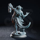 Rear-facing view of the same fish-headed aberration, revealing a scaled back, tattered layered cloth, a long reptilian tail sweeping behind, and an ornately carved stone plinth forming part of the scenic rocky base. Sculpt by DM Stash.