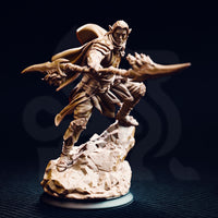 Photograph of the printed Reyvith the Valiant miniature, unpainted grey resin, showing the dual-blade lunging pose in close detail — elven features, chainmail and leather armour, sweeping cloak, and scenic stone base clearly visible at 32mm scale