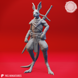 Unpainted grey resin render of the Aussie-Folk 09 kangaroo-folk warrior miniature, shown from a three-quarter front angle. The figure stands upright with digitigrade legs and a heavy tail, wearing shoulder armour, wrapped forearms, a neck scarf, and a belted sash. Two swords are crossed on the back with hilts visible above the shoulders.