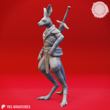 Side profile view of the unpainted grey Aussie-Folk 09 kangaroo-folk warrior miniature, showing the crossed sword hilts above the shoulder, chest harness, loose neck scarf, and the figure's strong digitigrade stance with powerful hindquarters and tail.