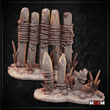 Digital render of two rustic wooden fence terrain pieces by Mia Kay M3DM — a large five-stake palisade section and a small two-stake broken section, both with rope bindings and scrubby ground detail, shown in warm grey resin tones against a dark background