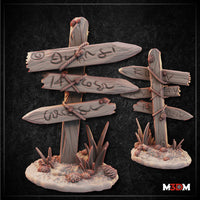Digital render of two rustic wooden signpost terrain pieces by Mia Kay M3DM. The larger post has four rope-bound planks carved with glyphs and numerals pointing in multiple directions; the smaller has three boards. Both sit on oval bases with desert scrub grass and bulbous plants. Unpainted resin colourway.