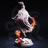 Rear view of the unpainted Selrina resin miniature showing the heavily textured woven cloak, windswept hair tipped with flame, sword pommel visible at her side, and sculpted orange flame effects rising from the rubble-strewn base