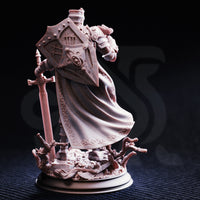 Photograph of printed unpainted grey resin Ser Ludwig miniature — rear three-quarter view highlighting the detailed kite shield with cross and devotional engravings strapped to his back, scrollwork-trimmed cloak sweeping dramatically behind him, and the scenic rubble and root base