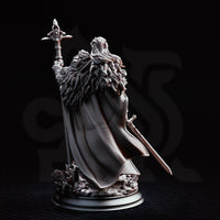 Silvia the Tearless war priestess miniature by DM Stash, holding a tall ornate cross-topped staff aloft, longsword at her side, wearing layered fur pauldrons and a billowing cloak, standing on a shattered rocky base.
