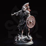 Skjolnir Ulvar by DM Stash — wolf-skulled barbarian in a coiled ready stance, mace raised at shoulder height, round wooden shield at his side. Layered fur and scale armour with wrapped leather leg bindings. Decorative base with wild scrub and scattered debris.