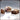 Digital render of the Novaminis Supplies 03 scatter terrain set showing three unpainted resin pieces: a stack of three stitched canvas sacks, a wooden crate filled with bread loaves, and a small domed wooden chest with a handle, all in a warm brown clay colour