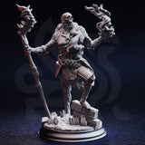 Front view of an unpainted grey resin miniature render — a skeletal undead sorcerer striding forward, one hand gripping a tall spiked bone staff, the other raised with necrotic soul-flame curling from the fingers. The figure has exposed skull, rotting sinew-wrapped musculature, tattered chainmail, and stands on a cracked stone rubble scenic base.