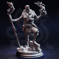 Front view of an unpainted grey resin miniature render — a skeletal undead sorcerer striding forward, one hand gripping a tall spiked bone staff, the other raised with necrotic soul-flame curling from the fingers. The figure has exposed skull, rotting sinew-wrapped musculature, tattered chainmail, and stands on a cracked stone rubble scenic base.