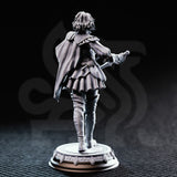 Photograph of printed grey resin Victoria Asquith Lady Duellist miniature by DM Stash — rear three-quarter view, unpainted, showing the full sweep of the cape, back detail of the tiered skirt, belt and scabbard, tall boots with stitching detail, and the reverse of the ornate circular base.