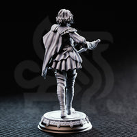 Photograph of printed grey resin Victoria Asquith Lady Duellist miniature by DM Stash — rear three-quarter view, unpainted, showing the full sweep of the cape, back detail of the tiered skirt, belt and scabbard, tall boots with stitching detail, and the reverse of the ornate circular base.