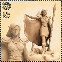 Digital render of an unpainted elven female miniature named Mia Kay, shown from two angles — full body and close-up bust. She wears layered pauldron armour with scrollwork detailing, holds a tall spear in one hand and draws back a dramatically billowing cloak with the other. Her eyes are closed in serene concentration. She stands on a naturalistic rocky base. Sculpted by Mia Kay M3DM.