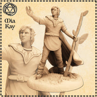 Unpainted resin render of a half-elf druid miniature by Mia Kay M3DM, standing on a rocky natural base. He holds a tall wooden staff adorned with leaves in one hand and raises the other skyward. He wears layered robes with a broad leather belt, fur-trimmed boots, and a flowing cape. A close-up inset shows his focused expression and pointed ears. 32mm scale.