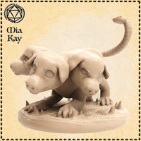 Digital render of Baby Cerberus miniature by Mia Kay M3DM — a three-headed hellhound pup sculpted in sandy beige resin tone, shown unpainted. Three blocky puppy heads cluster together on a stout body, with wide blank eyes and snub noses. A serpent-headed tail curls upward behind the figure. A row of dorsal spines runs down the back. The sculpted round base features stone fragments and emerging spines. Shown against a parchment-toned decorative border with the artist logo in the top left corner.
