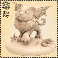 Unpainted resin render of a baby manticore miniature by Mia Kay M3DM — a stocky winged lion cub with a curled scorpion tail, in a low prowling pose with one clawed paw extended over a rocky scenic base scattered with broken stone shards, printed at 32mm scale