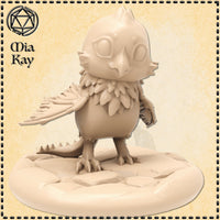 Unpainted resin miniature of a chibi baby cockatrice — round feathered body, owl-like skull face with wide blank eyes, layered chest feathers, one wing half-spread, scaled tail with small dorsal spines, standing on a sculpted rocky base. Digital render in a warm sandy tone by Mia Kay M3DM.